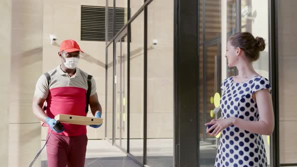 African Courier in Mask Deliver Pizza to Young Woman at Business Center Entrance