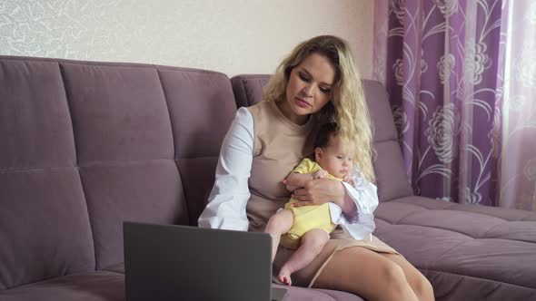 Busy Young Mother Works on Laptop Holding Baby Girl on Knees