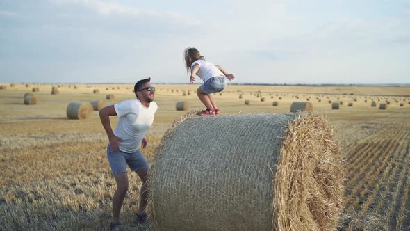 Little Daughter Jumping on Father's Arms From a Big Haycock in Field