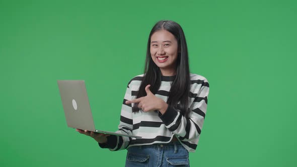 A Smiling Asian Woman Pointing To Computer While Standing In Front Of Green Screen Background