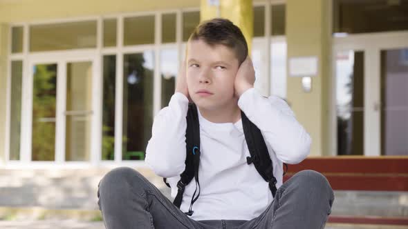 A Caucasian Teenage Boy Hears a Painful Noise and Covers His Ears As He Sits in Front of School