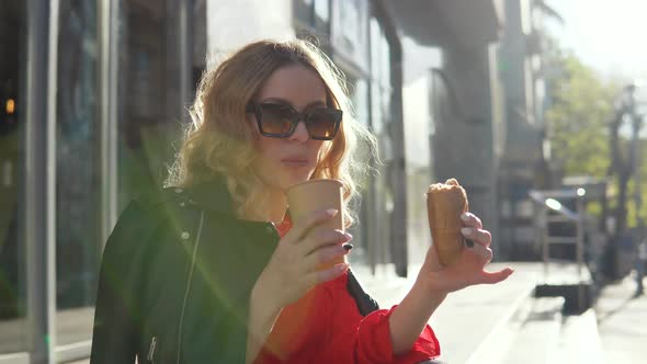 Stylish Beauty Drinks Coffee and Eats a Croissant Sitting on the Stairs at the Entrance to a Modern