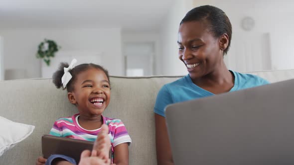 Happy african american mother and daughter sitting on sofa using digital tablet, laptop and laughing