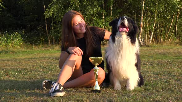 A Woman and a Border Collie Sit in a Meadow with a Trophy, the Woman Shows the Trophy To the Dog