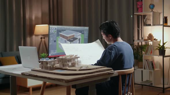 Male Engineer Opening And Looking At The Blueprint While Designing House On A Desktop At Home