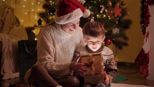 Happy Mother Hugging Her Son Unwrapping Christmas Gifts and Present in Living Room