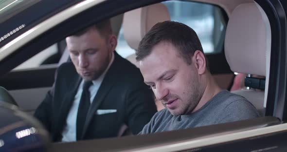 Close-up of Caucasian Young Man Sitting in Car Salon with Blurred Dealer at the Background