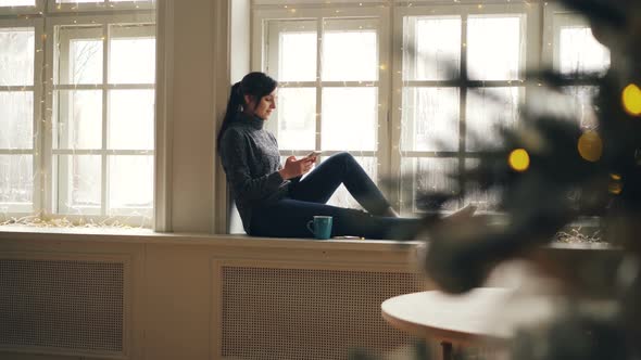 Smiling Young Lady Is Texting Friends on Christmas Day Using Smartphone Sitting on Window-sill