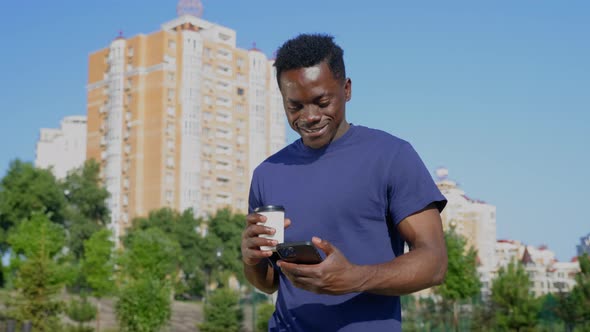 Smiling Afroamerican Man Walks Street Uses Mobile Phone Holding Cup with Coffee