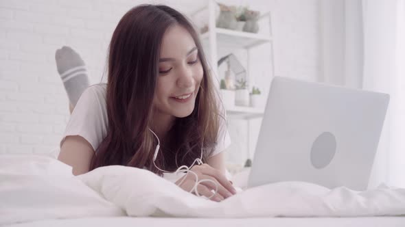 Asian woman using computer or laptop and listening music while lying on the bed.