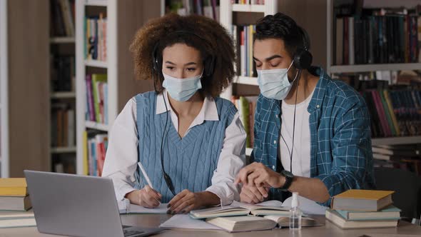 Two Students in Medical Masks Sit at Desk in University Library Listen to Teacher in Headphones on