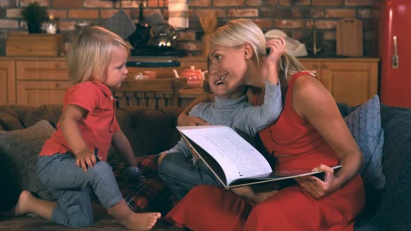 A Young Mother Sits on a Sofa with Two Children, Boys, a Book in Her Hands, an Older Brother Says