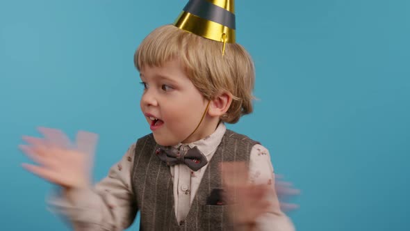 Smiling Little Birthday Boy in Cone Hat Applauding on Blue Wall Background