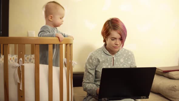 Woman is Sitting on Sofa with Laptop on Her Knees Next to Bed with Baby Inside and Talking By Video