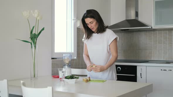 Young woman in white t-shirt prepares green smoothie in blender at home in the kitchen