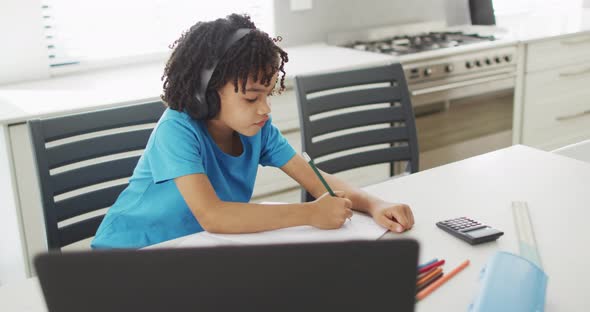 Happy biracial boy sitting at table in kitchen using laptop and doing homework