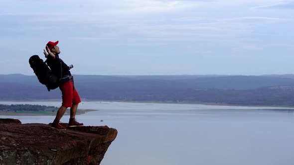 cheerful hiker man gesture raised arms on the edge of cliff, on a top of the rock mountain