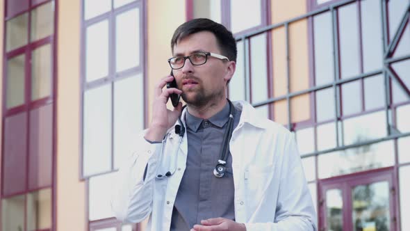 A Young Male Doctor Wears Glasses and Uses a Smartphone Outside the Entrance to the Hospital