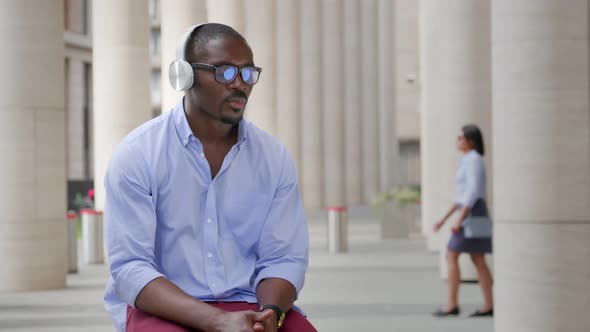 African Businessman in Financial District Listening Audio Songs in Headphones Sitting Outside