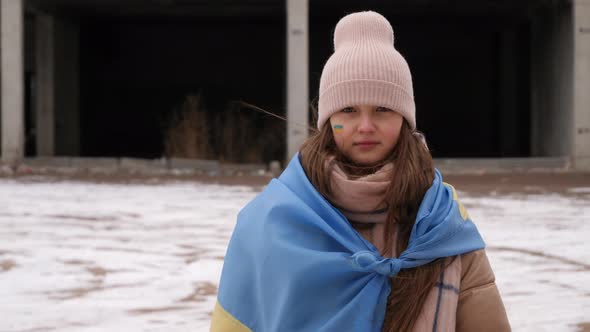 a girl wearing in the Ukrainian flag looks sadly at the camera against the background of destroyed h