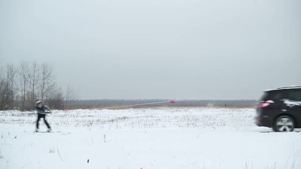 A car passes through the frame, followed by a snowboarder holding onto a rope