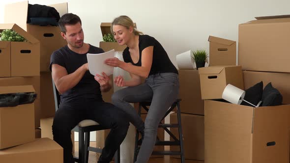 A Happy Moving Couple Sits on Chairs in an Empty Apartment and Reads a Bunch of Papers