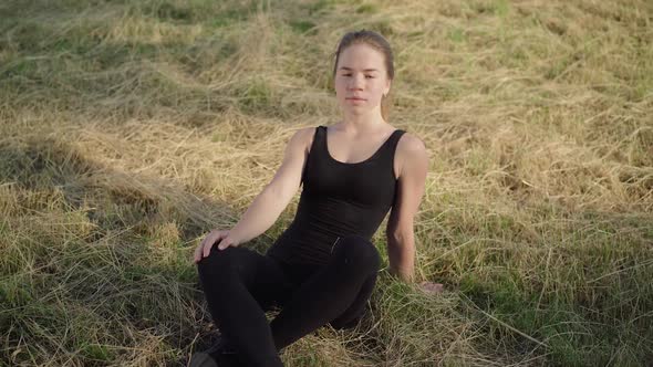 Relaxed Young Woman in Sportswear Sitting on Autumn Grass and Looking at Camera. Portrait of