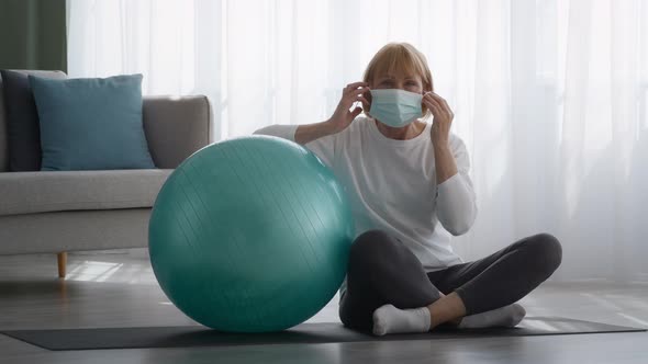 Senior Lady Putting On Mask Sitting Near Fitball At Home