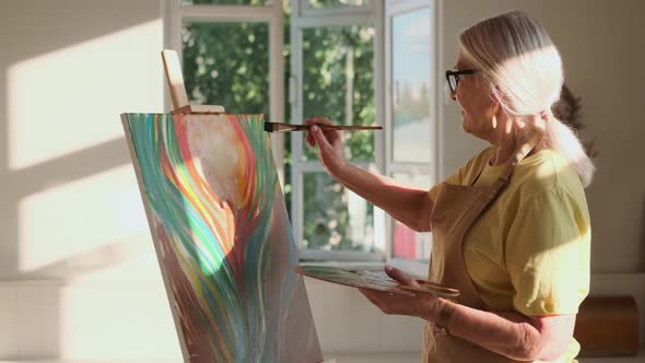 Elderly Woman Artist Paints a Picture on Canvas While Standing in a Sunny Workshop