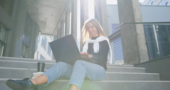 Girl in Casual Clothes Sitting on Building's Steps and Working on Laptop with Satisfied face