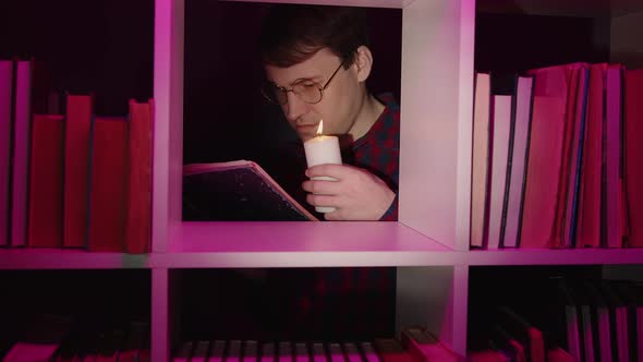 Man in Glasses with Candle Reading Book Standing Behind Bookshelf