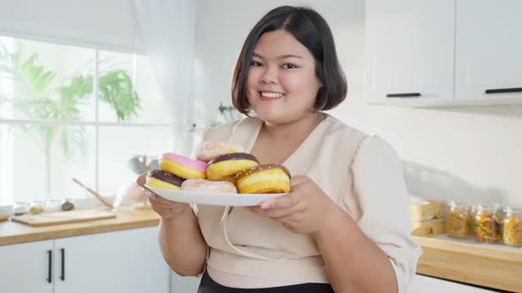 Portrait of Asian oversize big women holding donut on plate enjoy unhealthy foods in kitchen.