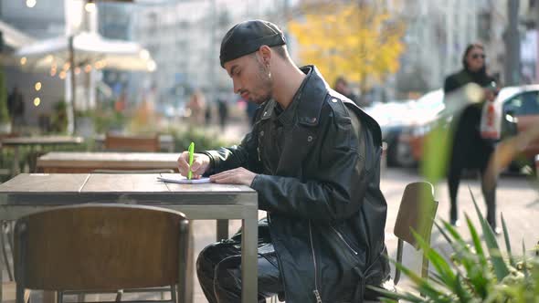 Side View Thoughtful Young Gay Man Sitting at Table in Sidewalk Cafe Writing with Pen