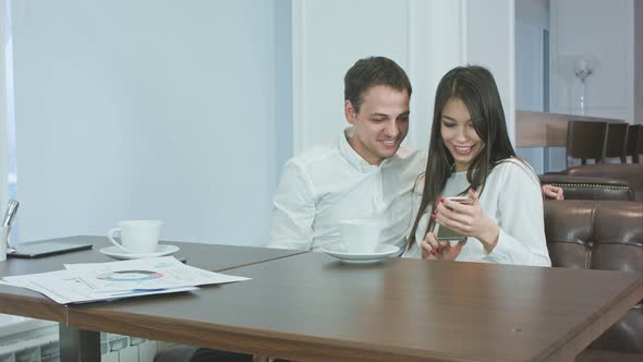 Two Office Coworkers in a Cafe Looking at Their Photos on the Phone and Laughing