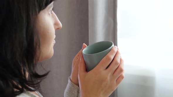 The Girl Is Holding a Cup of Hot Tea or Coffee. A Woman Enjoys a Hot Drink While Looking Out the