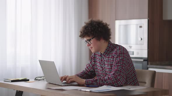 A Curly Man with a Serious Look Works at a Laptop Sitting in a Modern Kitchen