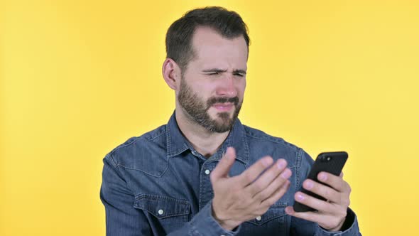 Beard Young Man Reacting To Loss on Smartphone, Yellow Background