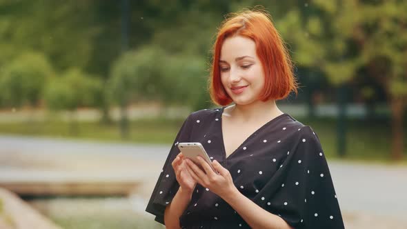 Beautiful Focused Redhead Caucasian Girl Teenage User Woman with Short Hair Stand in City Park Hold