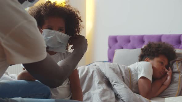 African Father Putting Safety Mask on Little Daughter with Sick Son Lying in Bed on Background