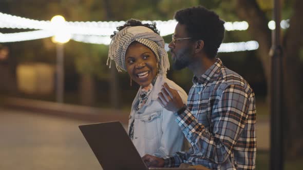 Black Man and Woman Using Laptop and Chatting in Park in Evening