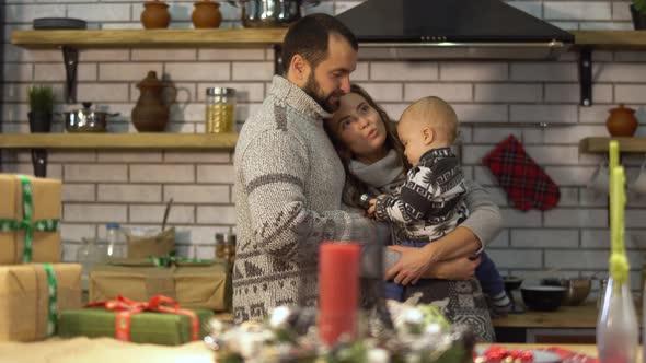 Bearded Father in Warm Sweater Playing with Baby Little Son in Mother Arms in the Kitchen