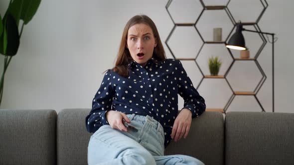 Closeup Portrait of a Young Woman Sitting on a Sofa Watching TV Holding a Remote Control Shocked By