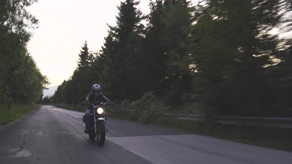 Male Biker Riding Motorcycle on Forest Road in the Evening with Turned Headlight