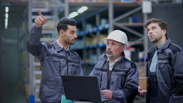 Senior Expert Man in Hard Hat Showing Logistics Plan on Laptop to Employees Talking Pointing Up at