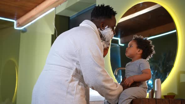 Young African American Positive Father Playing with Toddler Son Applying Shave Foam in Bathroom