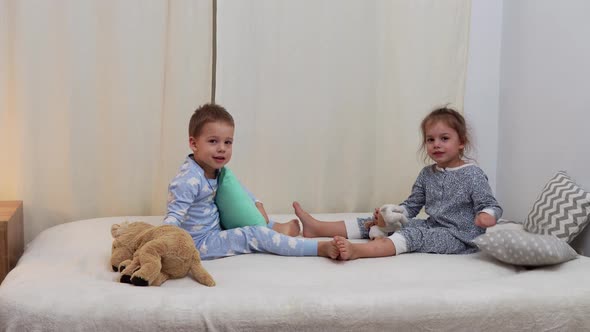 Two Smiling Preschool Toddler Children In Pajamas Playing With Teddy Bear on Bed