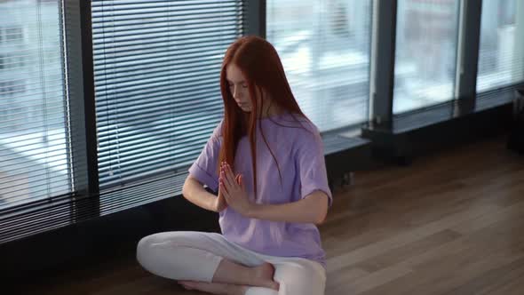 Young redhead woman meditating practicing breathing yoga exercise, sitting on floor near window