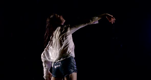 Dark-haired Girl in White Shirt and Denim Shorts, She Stands on a Black Background in Heavy Rain