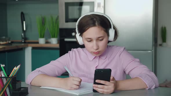 The Schoolgirl with Headphones Teaches Lessons While at Home Sitting in the Kitchen at the Table