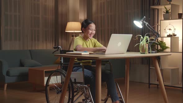 Asian Kid Girl Sitting In A Wheelchair While Using Laptop Computer And Celebrating At Home